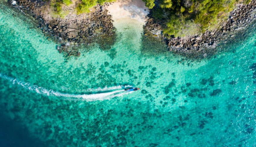 View from above, aerial view of a traditional long-tail boat sailing near a stunning barrier reef with a beautiful small beach bathed by a transparent and turquoise sea. Phi Phi Island, Thailand.